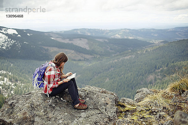 Frau benutzt Tablet-Computer  während sie auf dem Gipfel eines Berges vor bewölktem Himmel sitzt