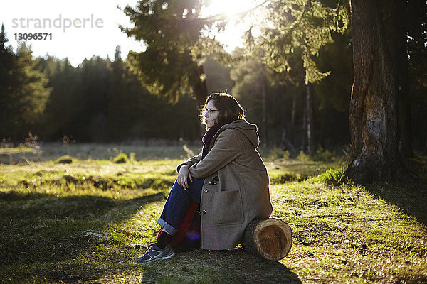 Seitenansicht einer nachdenklichen Frau  die wegschaut  während sie auf einem Baumstamm im Wald sitzt
