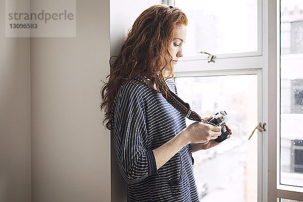 Frau benutzt Digitalkamera  während sie zu Hause am Fenster steht