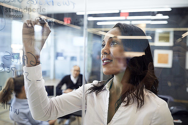 Geschäftsfrau schreibt Plan auf Glas im Sitzungssaal mit Kollegen im Hintergrund