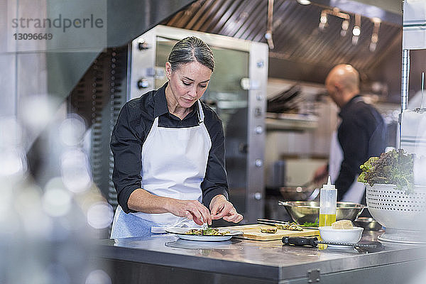 Köchin beim Garnieren von Speisen  während eine Mitarbeiterin im Hintergrund in der Restaurantküche arbeitet
