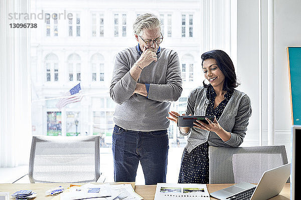 Geschäftsfrau zeigt einem leitenden Geschäftsmann einen Tablet-Computer  während sie im Kreativbüro am Fenster steht
