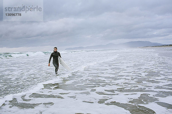 Mann mit Surfbrett schaut weg  während er im Meer gegen bewölkten Himmel läuft
