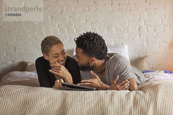 Glückliches Paar unterhält sich  während es zu Hause am Bett einen Tablet-Computer benutzt