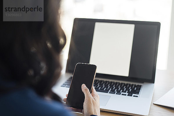 Ausgeschnittenes Bild einer Geschäftsfrau  die ein Mobiltelefon benutzt  während sie im Büro an einem Laptop-Computer sitzt