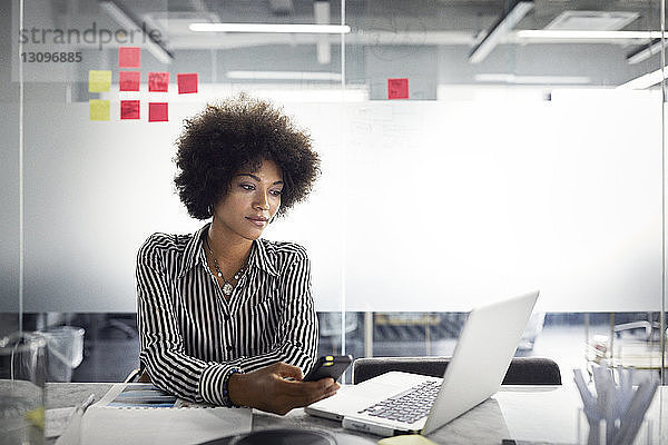 Geschäftsfrau schaut auf Laptop-Computer  während sie ein Smartphone im Büro hält
