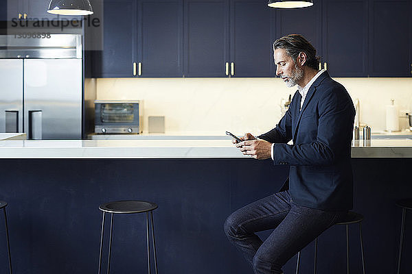 Geschäftsmann benutzt ein Smartphone  während er auf einem Hocker an der Küchentheke im Kreativbüro sitzt