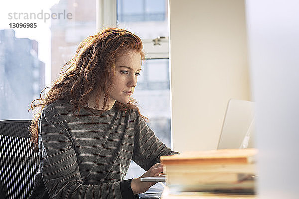 Zuversichtliche Frau benutzt Laptop-Computer  während sie zu Hause am Fenster sitzt