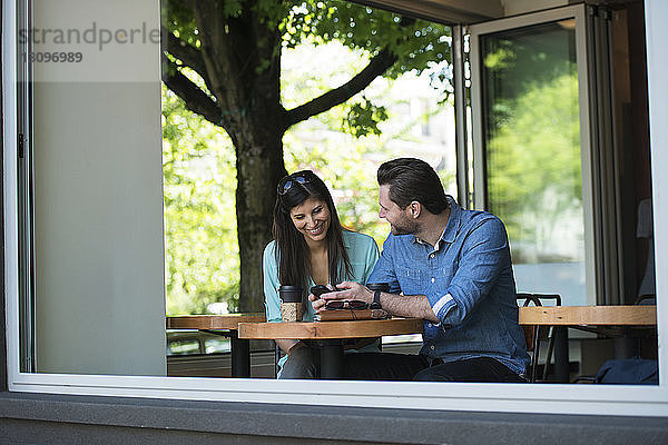 Glückliches Paar  das im Café sitzend durchs Fenster auf sein Handy schaut