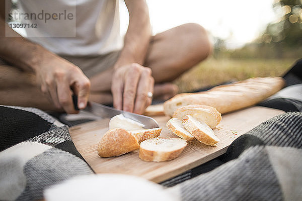 Niedriger Teil eines Mannes schneidet Brot  während er auf einer Picknickdecke sitzt