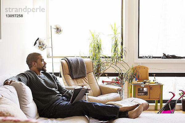 Mann mit Tablet-Computer schaut weg  während er zu Hause auf dem Sofa sitzt