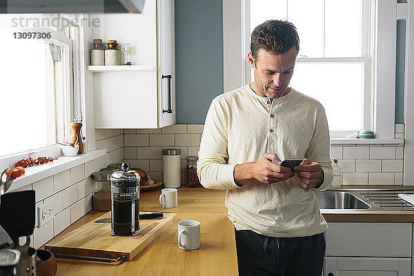 Mann benutzt Smartphone  während er sich zu Hause an die Küchentheke lehnt