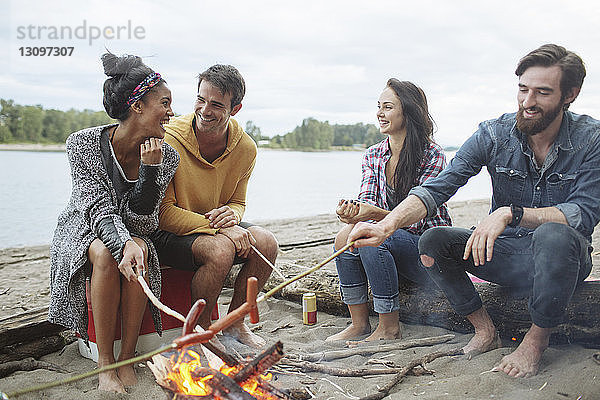 Glückliche Freunde braten Würstchen  während sie am Lagerfeuer am Fluss sitzen