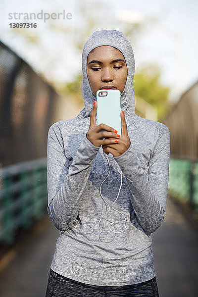 Frau trägt Kapuzenhemd  während sie ein Smartphone auf einem Fußweg benutzt