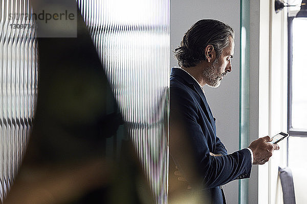 Geschäftsmann benutzt Smartphone  während er sich im Büro an eine Glaswand lehnt