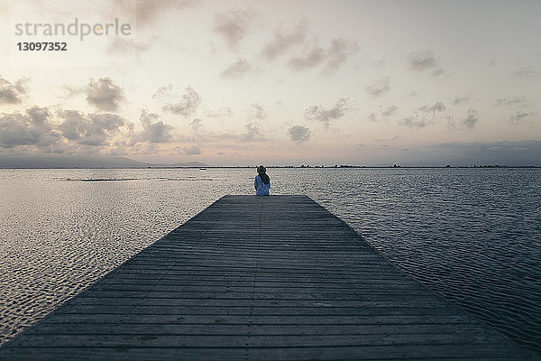 Rückansicht einer Frau  die bei Sonnenuntergang auf dem Pier über dem Meer gegen den Himmel sitzt