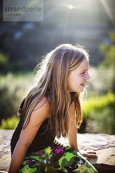 Seitenansicht eines lächelnden Mädchens auf einer Stützmauer bei sonnigem Wetter