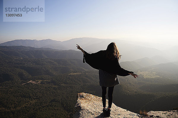 Rückansicht einer Frau  die gestikulierend auf einer Klippe vor Bergen steht