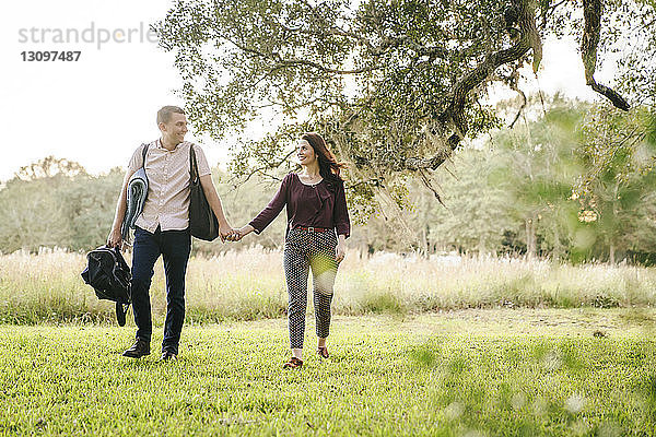 Lächelndes Paar hält sich beim Spaziergang auf dem Grasfeld im Park an den Händen