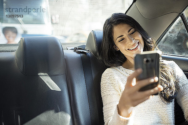 Glückliche junge Frau  die sich im Taxi durch ein Smartphone selbstständig macht