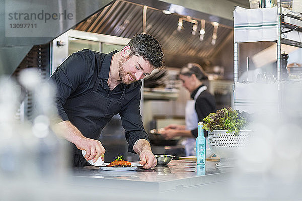 Chefkoch beim Garnieren von Essen  während eine weibliche Mitarbeiterin im Hintergrund im Restaurant arbeitet