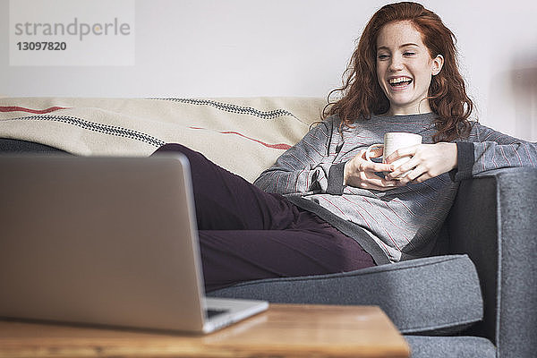 Fröhliche Frau mit Tasse schaut auf Laptop-Computer  während sie zu Hause auf dem Sofa sitzt