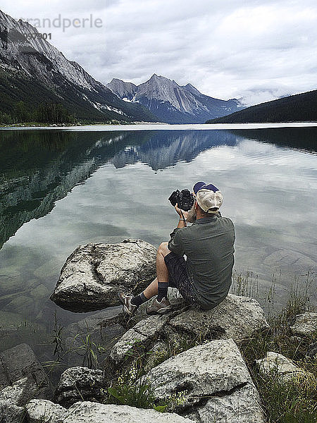 Mann sitzt auf Fels und fotografiert See mit Kamera