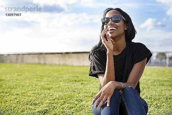 Fröhliche Frau mit Sonnenbrille auf einem Grasfeld im Park sitzend