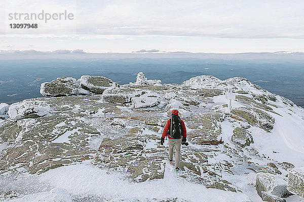 Rückansicht eines Rucksacktouristen mit Kamera beim Gehen auf schneebedeckten Felsen