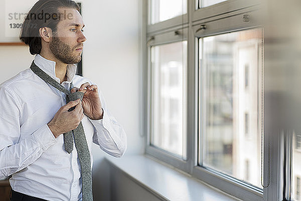 Nachdenklicher Mann schaut durch das Fenster und bindet Krawatte zu Hause