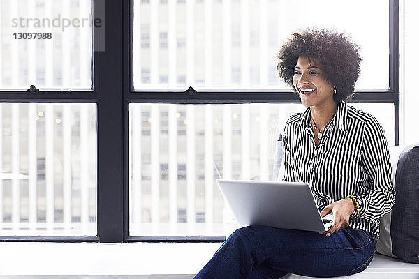 Fröhliche Geschäftsfrau benutzt Laptop-Computer  während sie im Büro am Fenster sitzt