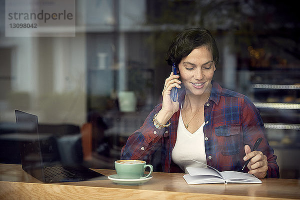 Frau spricht am Smartphone  während sie im Café sitzt und durch das Fenster gesehen wird
