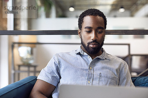 Junger selbstbewusster Geschäftsmann mit Laptop  während er im Kreativbüro auf dem Sofa sitzt