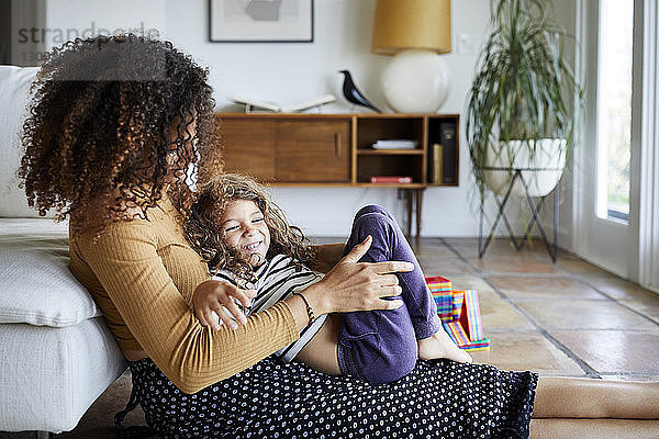 Fröhliche Mutter spielt mit ihrer Tochter  während sie zu Hause auf dem Boden am Sofa sitzt