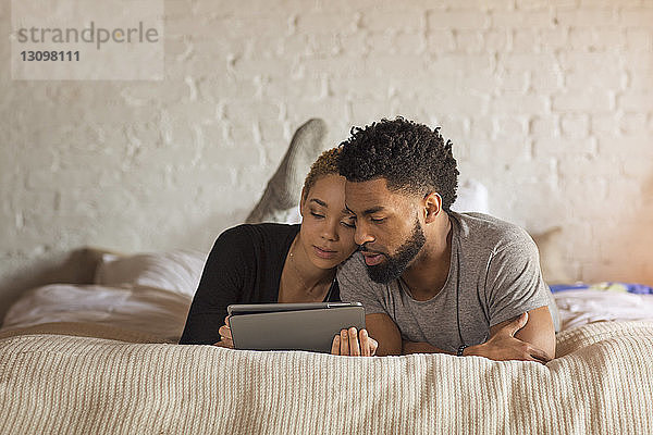 Multi-ethnisches Paar benutzt Tablet-Computer  während es zu Hause im Bett liegt