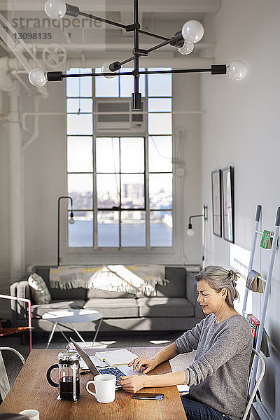 Konzentrierte reife Frau mit Laptop bei der Arbeit am Tisch im Wohnzimmer