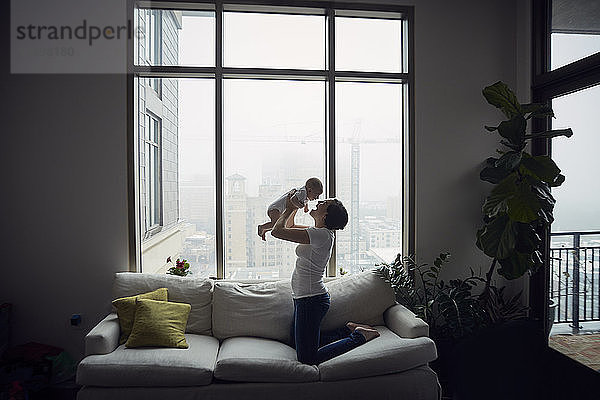 Seitenansicht einer glücklichen Mutter  die mit ihrem Sohn spielt  während sie zu Hause auf dem Sofa am Fenster kniet
