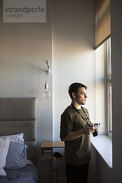 Mann mit Kamera  der durch ein Fenster schaut  während er zu Hause im Schlafzimmer steht