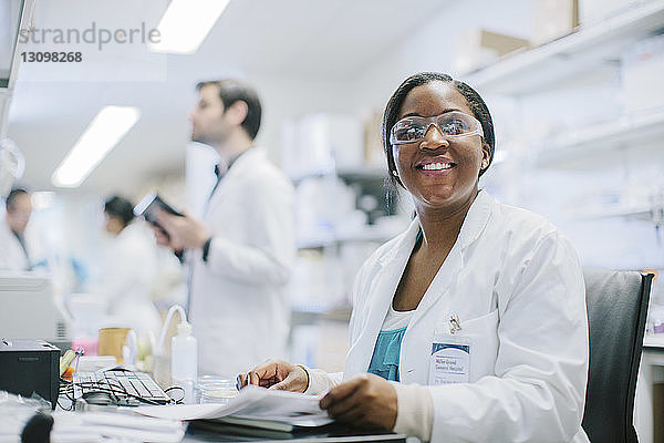 Porträt einer lächelnden Ärztin  die am Schreibtisch sitzt  während ihre Kollegen im Behandlungszimmer arbeiten