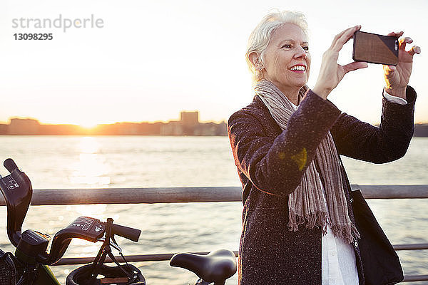 Glückliche ältere Frau fotografiert bei Sonnenuntergang am Meer