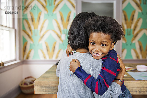 Porträt eines glücklichen Jungen  der seine Mutter umarmt  während er zu Hause Buntstifte hält