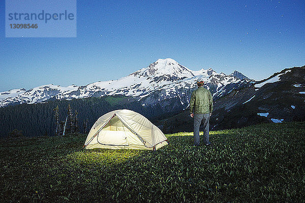 Rückansicht eines Mannes  der bei beleuchtetem Zelt auf einem Grasfeld vor einem Berg in der Dämmerung steht