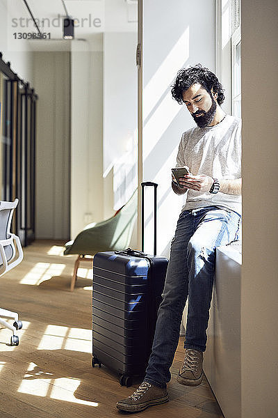 Geschäftsmann in voller Länge  der ein Smartphone benutzt  während er im Kreativbüro auf dem Fensterbrett sitzt