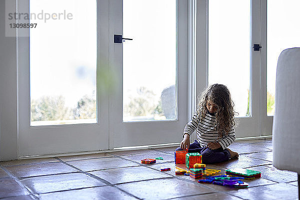 Mädchen spielt mit bunten Spielzeugbausteinen  während sie zu Hause auf dem Boden kniet