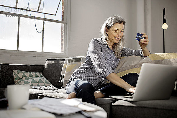 Reife Frau hält Debitkarte  während sie den Laptop auf dem Sofa benutzt