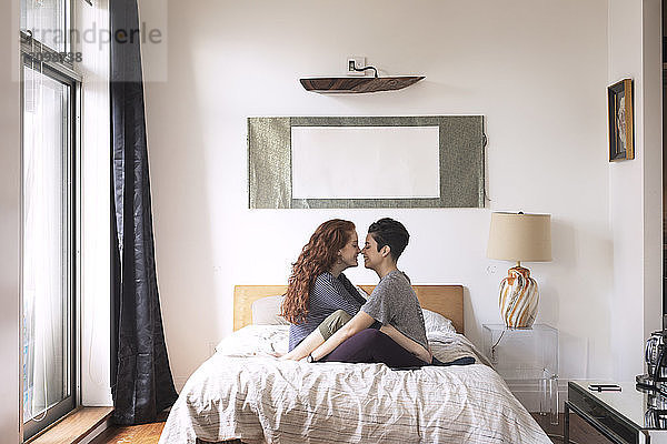 Seitenansicht von romantischen Lesben  die sich die Nasen reiben  während sie auf dem Bett sitzen
