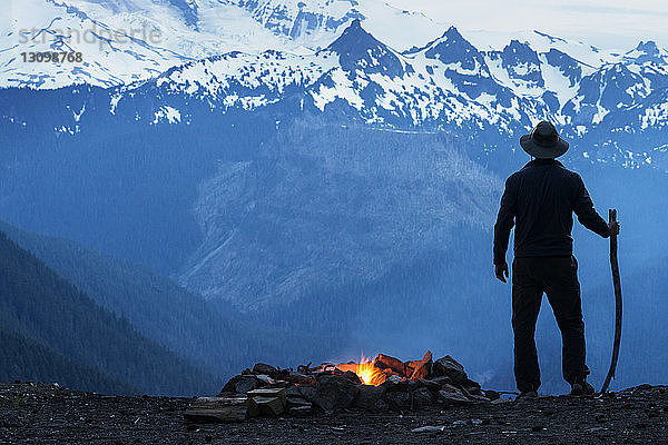 Mann hält Stock am Lagerfeuer gegen schneebedeckte Berge