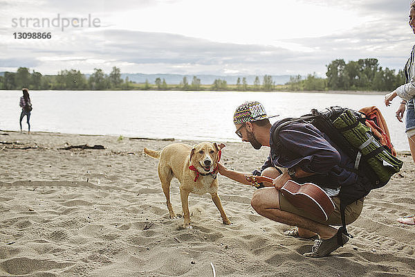 Seitenansicht eines Mannes  der einen Hund streichelt  während er auf Sand am Flussufer kauert