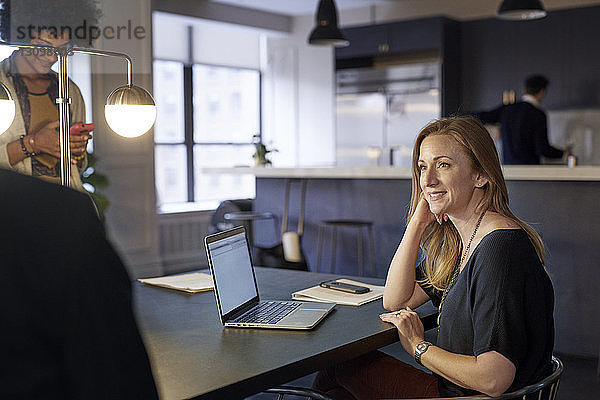 Glückliche Geschäftsfrau schaut weg  während eine Kollegin ein Smartphone im Kreativbüro benutzt