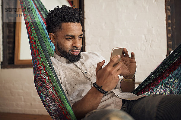 Mann benutzt Smartphone  während er zu Hause auf der Hängematte sitzt